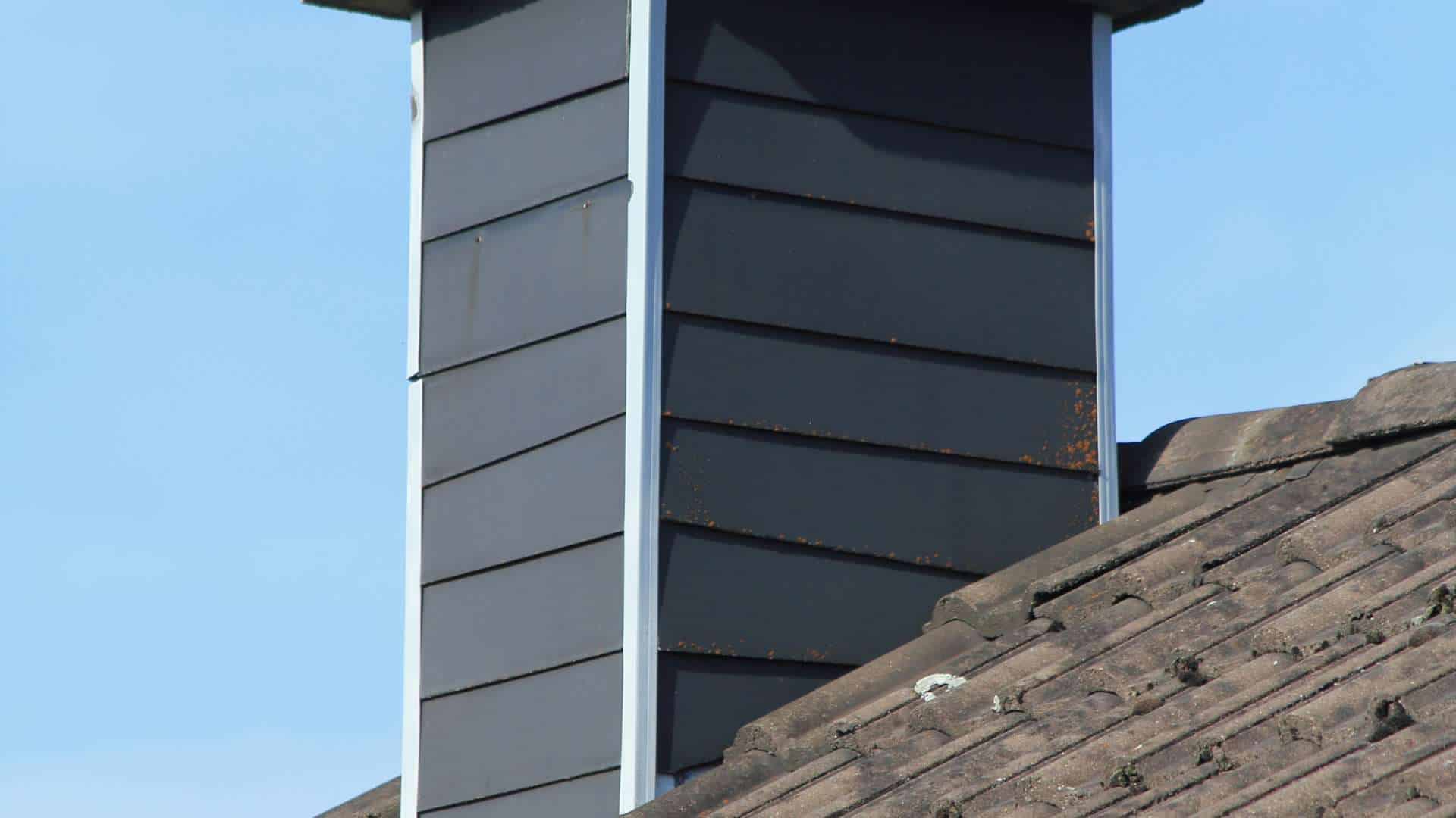 A close-up view showcases a chimney with dark gray siding on a slanted rooftop, expertly installed by the Chimney Company. The roof features brown shingles, and the sky is clear and blue in the background.