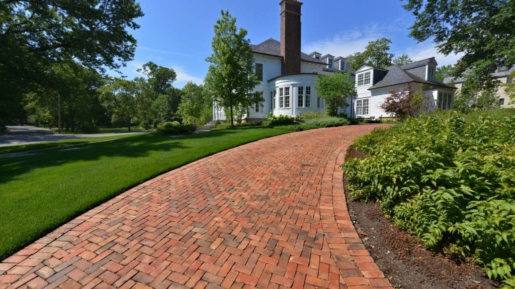 A red brick driveway curves towards a large, elegant house surrounded by lush green grass and tall trees. The prominent chimney, expertly maintained by a local chimney company, complements the large windows and well-kept garden. The sky is clear and blue.