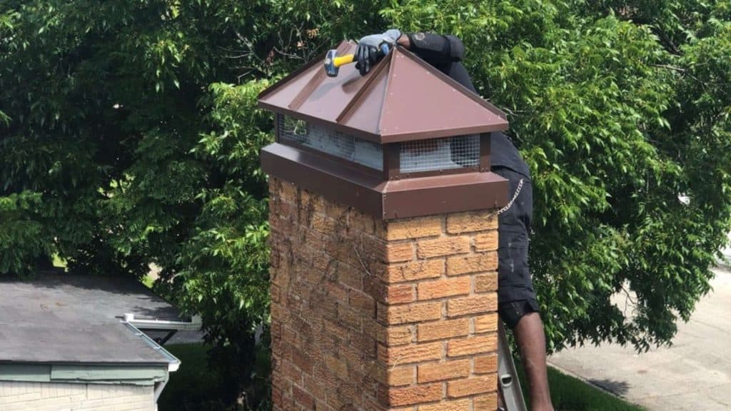 A person in protective gloves and clothing climbs a ladder to repair the top of a brick chimney. They expertly fit chimney caps, surrounded by green leafy trees and rooftops, showcasing the skill that only comes from working with a professional chimney company.