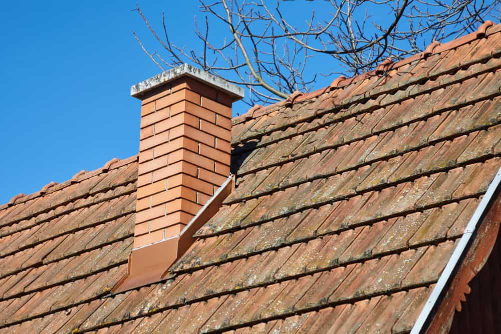 A red brick chimney, recently serviced by a chimney sweep, stands on a roof covered with weathered, brownish-red tiles. Bare branches stretch over a clear blue sky in the background.