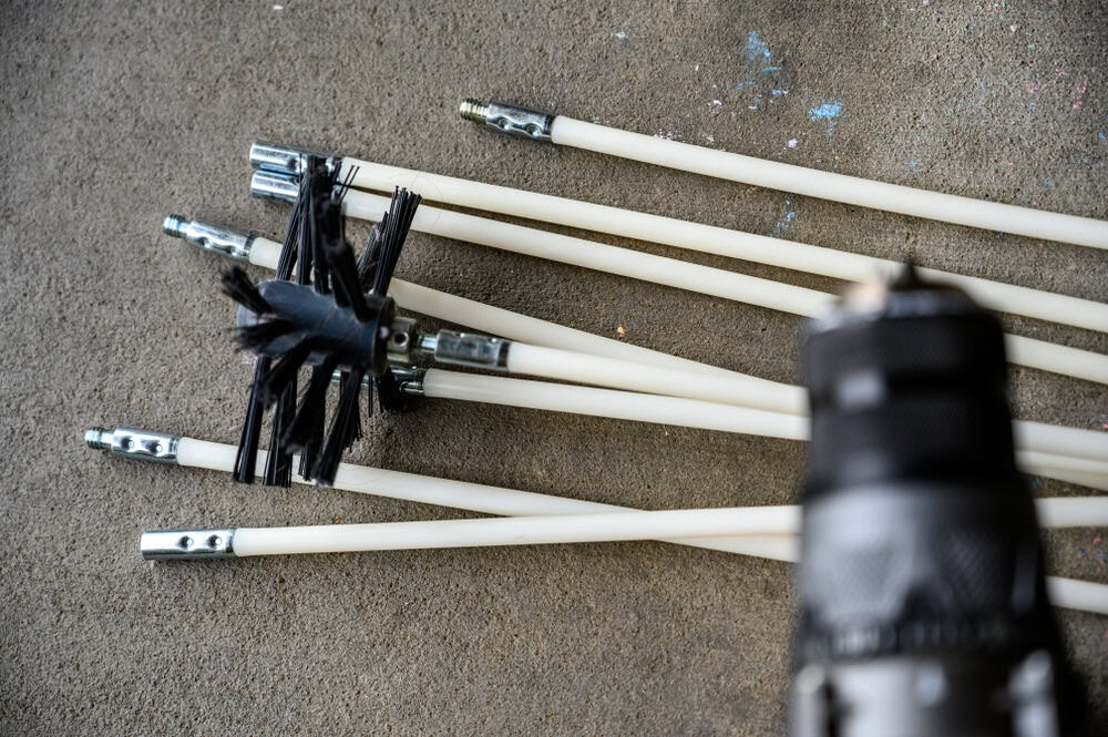A collection of white rods with metal ends, one wrapped in black tape, rests on a concrete surface, suggesting chimney sweep equipment. In the foreground is a blurred black cylindrical object, possibly a tool integral to chimney repair work.